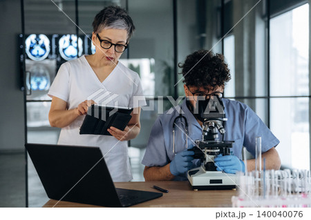 Female healthcare professional observes male colleague using microscope in laboratory, with digital tablet and laptop on table, medical images displayed in background 140040076