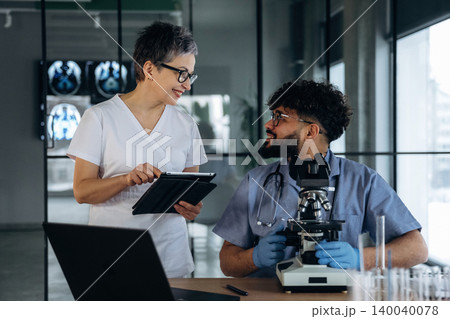 Healthcare professionals in laboratory discussing results, one holding a tablet and the other seated with a microscope, glassware and medical images in the background Healthcare professionals in laboratory discussing results, one holding a tablet and the other seated with a microscope, glassware and medical images in the background 140040078