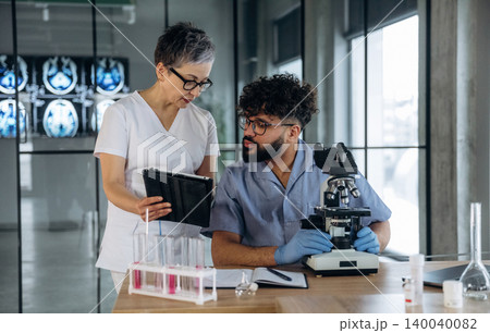 Female healthcare professional in white scrubs discusses findings with male laboratory technician holding microscope in modern laboratory with glass walls and equipment Female healthcare professional in white scrubs discusses findings with male laboratory technician holding microscope in modern laboratory with glass walls and equipment 140040082