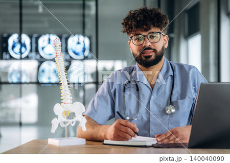 Male healthcare professional in blue scrubs with stethoscope sits at desk, writing notes beside anatomical spine model and laptop, medical imaging displayed in background 140040090