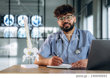Male healthcare professional in blue scrubs with stethoscope sits at desk, writing notes beside a spinal model and laptop, medical images displayed in background Male healthcare professional in blue scrubs with stethoscope sits at desk, writing notes beside a spinal model and laptop, medical images displayed in background 140040091