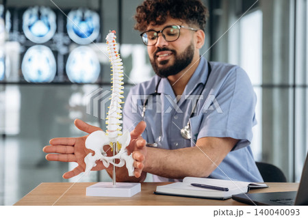 Male healthcare professional in scrubs explains spinal anatomy using a model on a desk, with medical imaging displayed in the background Male healthcare professional in scrubs explains spinal anatomy using a model on a desk, with medical imaging displayed in the background 140040093