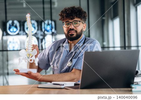 Male healthcare professional in scrubs holds a spinal model while sitting at a desk with a laptop and medical images displayed in the background Male healthcare professional in scrubs holds a spinal model while sitting at a desk with a laptop and medical images displayed in the background 140040096