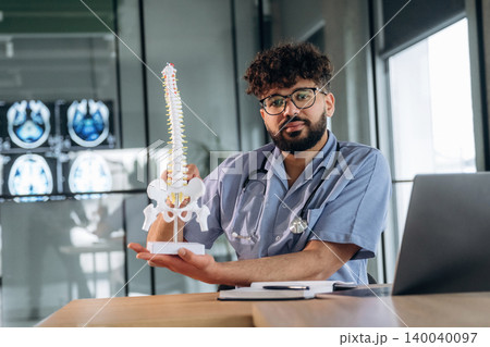 Male healthcare professional in scrubs holding a spinal model while seated at a desk with medical imaging displayed in the background Male healthcare professional in scrubs holding a spinal model while seated at a desk with medical imaging displayed in the background 140040097