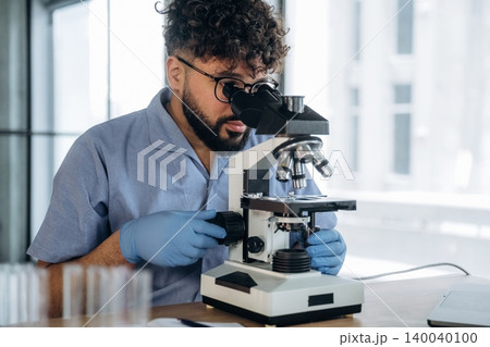 Male scientist wearing blue lab coat and gloves examines samples under a microscope in a bright laboratory with glass test tubes on the table Male scientist wearing blue lab coat and gloves examines samples under a microscope in a bright laboratory with glass test tubes on the table 140040100