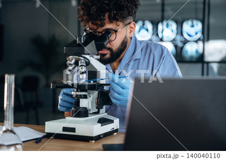 Male scientist in blue lab coat examines specimen under microscope in modern laboratory with medical imaging screens visible in the background 140040110