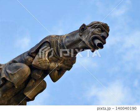 Weathered carved stone gargoyle jutting from roof edge showcasing open mouth and expressive features Weathered carved stone gargoyle jutting from roof edge showcasing open mouth and expressive features 140040190