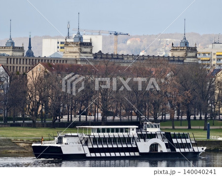 Passenger ferry docked beside treelined bank. Calm reflective water with cityscape in background Passenger ferry docked beside treelined bank. Calm reflective water with cityscape in background 140040241