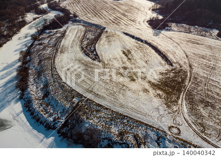 Drone shot of patchy snowy fields, revealing groovy furrows and tire marks. A frozen river snuggles alongside, a chill winter scene Drone shot of patchy snowy fields, revealing groovy furrows and tire marks. A frozen river snuggles alongside, a chill winter scene 140040342