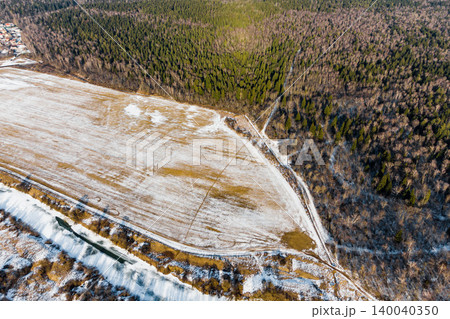 Aerial view reveals a meandering river or stream, partially frozen, separating a vast, snow-dusted field from a dense, wintry forest. A serene, chilly landscape 140040350