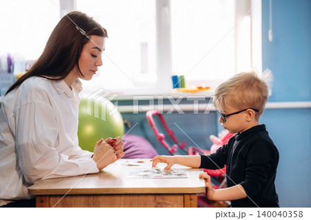 Preschool child with teacher engaged in developmental activity at a wooden table in a colorful classroom with educational materials and toys visible in the background Preschool child with teacher engaged in developmental activity at a wooden table in a colorful classroom with educational materials and toys visible in the background 140040358