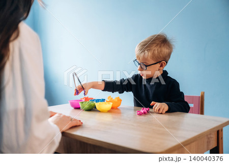 Preschool child with glasses using tongs to pick up colorful play food during developmental activity with teacher in a bright classroom setting Preschool child with glasses using tongs to pick up colorful play food during developmental activity with teacher in a bright classroom setting 140040376