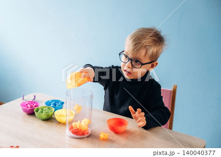 Preschool child with glasses engaged in developmental play, placing colorful shapes into a transparent container on a wooden table with vibrant bowls in the background Preschool child with glasses engaged in developmental play, placing colorful shapes into a transparent container on a wooden table with vibrant bowls in the background 140040377