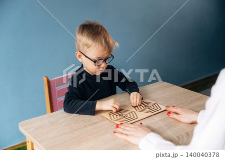Preschool child with glasses focused on geometric puzzle while teacher's hands guide him at a wooden table in a brightly colored classroom setting Preschool child with glasses focused on geometric puzzle while teacher's hands guide him at a wooden table in a brightly colored classroom setting 140040378