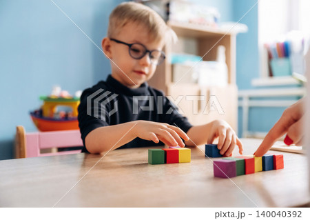 Preschool child with glasses engaged in color sorting activity with teacher's hand guiding on wooden table in bright classroom setting with educational materials visible Preschool child with glasses engaged in color sorting activity with teacher's hand guiding on wooden table in bright classroom setting with educational materials visible 140040392
