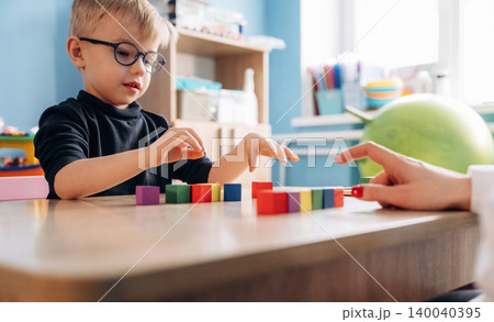 Preschool child with glasses engaged in developmental play with colorful building blocks at a wooden table alongside a teacher's hand in a bright classroom setting Preschool child with glasses engaged in developmental play with colorful building blocks at a wooden table alongside a teacher's hand in a bright classroom setting 140040395