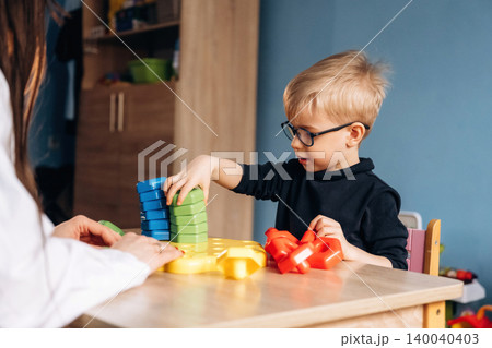 Preschool child with glasses stacking colorful building blocks with teacher's assistance at a wooden table in a bright classroom setting Preschool child with glasses stacking colorful building blocks with teacher's assistance at a wooden table in a bright classroom setting 140040403