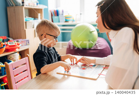 Preschool child with glasses engaged in a learning activity with female teacher at a colorful classroom table, educational materials and toys visible in the background Preschool child with glasses engaged in a learning activity with female teacher at a colorful classroom table, educational materials and toys visible in the background 140040435