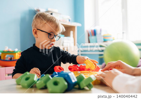 Preschool child with glasses playing with colorful building blocks on a table in a bright classroom, teacher's hand visible guiding the activity Preschool child with glasses playing with colorful building blocks on a table in a bright classroom, teacher's hand visible guiding the activity 140040442