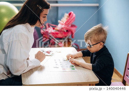 Preschool child with glasses engaged in a learning activity with a female teacher at a wooden table in a colorful classroom with toys in the background Preschool child with glasses engaged in a learning activity with a female teacher at a wooden table in a colorful classroom with toys in the background 140040446