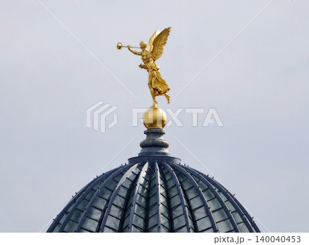 Lustrous gilded angel sculpture standing tall with trumpet raised against subdued sky backdrop Lustrous gilded angel sculpture standing tall with trumpet raised against subdued sky backdrop 140040453