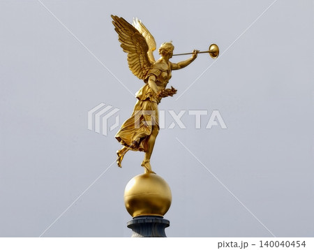 Radiant gilded angel on column. Luminous gold angel sculpture perched atop tall column in cityscape Radiant gilded angel on column. Luminous gold angel sculpture perched atop tall column in cityscape 140040454
