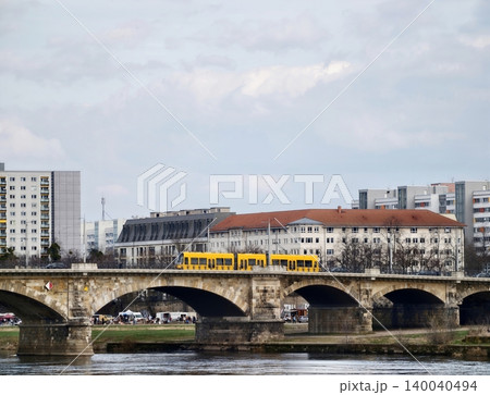 Yellow tram crossing stone bridge over river, modern apartments, overcast sky and waterfront reflections 140040494