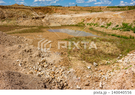 Open pit quarry with a reflective puddle, surrounded by dry, cracked earth and rubble under a blue sky with clouds 140040556