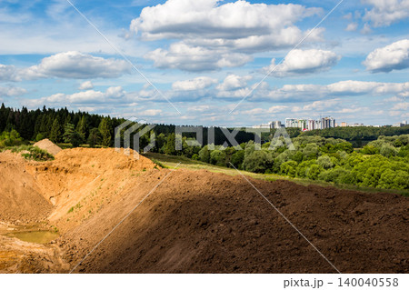 Expansive quarry site featuring rich sand and brown soil mounds, a deep excavation pit under a vibrant sky. Distant city buildings dot the horizon beyond green forests 140040558