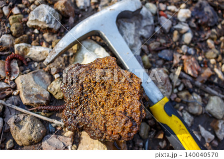 A distinctively textured, rugged rock specimen lies on rocky ground next to a geological hammer. This unique, bumpy mineral evokes exploration and natural discovery A distinctively textured, rugged rock specimen lies on rocky ground next to a geological hammer. This unique, bumpy mineral evokes exploration and natural discovery 140040570