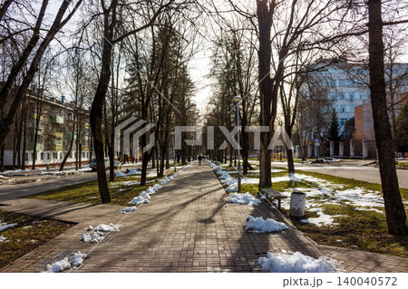 A long park alley in early spring, lined with bare trees casting shadows. Melting snow patches dot the ground, with a lone person walking in the distance along the pathway A long park alley in early spring, lined with bare trees casting shadows. Melting snow patches dot the ground, with a lone person walking in the distance along the pathway 140040572