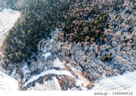 Aerial winter forest view. Evergreen and bare trees blanketed in snow near a frozen creek and snowy fields. Nature's frosty grip Aerial winter forest view. Evergreen and bare trees blanketed in snow near a frozen creek and snowy fields. Nature's frosty grip 140040618