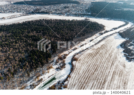Aerial winter vista featuring a winding, partially frozen river dividing a dark forest and a snowy field. Distant village barely visible Aerial winter vista featuring a winding, partially frozen river dividing a dark forest and a snowy field. Distant village barely visible 140040621