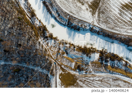 Icy river meandering through snow-covered winter terrain, flanked by a stark forest and frosty fields. A peaceful aerial vista of nature's chill 140040624