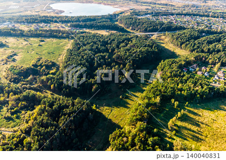 Aerial view of a sprawling green forest, interspersed with sunny fields and a quaint village. A tranquil lake shimmers in the distance, completing this serene natural panorama Aerial view of a sprawling green forest, interspersed with sunny fields and a quaint village. A tranquil lake shimmers in the distance, completing this serene natural panorama 140040831