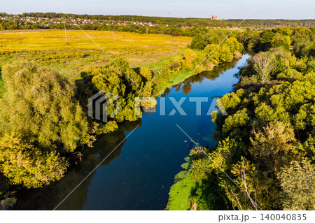 Winding river snaking through vibrant green and yellow foliage. Aerial shot captures tranquil waters reflecting the sky, surrounded by lush riverbanks and a distant rural landscape Winding river snaking through vibrant green and yellow foliage. Aerial shot captures tranquil waters reflecting the sky, surrounded by lush riverbanks and a distant rural landscape 140040835