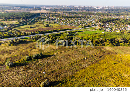 Motorway slicing through verdant terrain, bordered by dry fields and a quaint cottage settlement, with distant city structures peeking Motorway slicing through verdant terrain, bordered by dry fields and a quaint cottage settlement, with distant city structures peeking 140040836