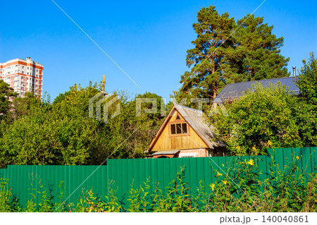 Charming rustic wooden cottage peeks over a vibrant green fence, nestled amidst lush trees. A tall apartment building hints at urban closeness under a clear sky 140040861