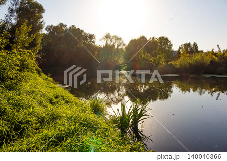 Lush green riverbank, tranquil waters reflect trees under golden sunlight. Perfect spot for a peaceful angling adventure Lush green riverbank, tranquil waters reflect trees under golden sunlight. Perfect spot for a peaceful angling adventure 140040866