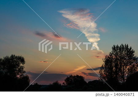 A dramatic, wispy cloud streaks across a vibrant twilight sky, blending warm sunset hues with cool blues above silhouetted trees 140040871