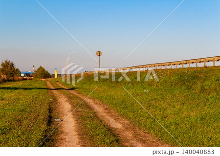 A rustic dirt track meanders beside a modern highway, with a protective guardrail delineating the edge. Distant streetlights and a gas station add hints of development A rustic dirt track meanders beside a modern highway, with a protective guardrail delineating the edge. Distant streetlights and a gas station add hints of development 140040883