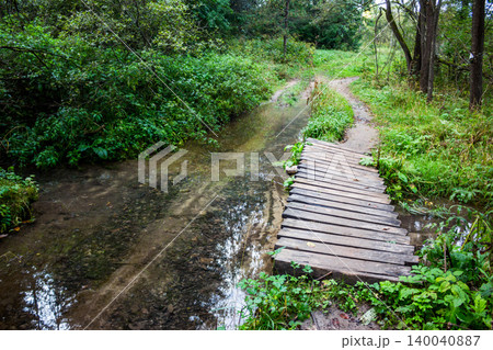 Weathered wooden footbridge crosses a clear brook, leading to a winding dirt path amidst lush greenery. Peaceful forest scene 140040887