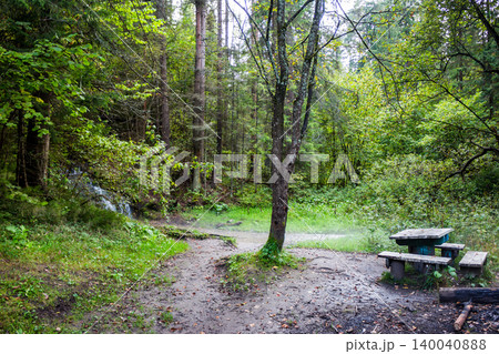 Quaint wooden picnic table and benches in a lush forest nook. A secluded spot for rest, surrounded by tall trees and the sound of a small stream Quaint wooden picnic table and benches in a lush forest nook. A secluded spot for rest, surrounded by tall trees and the sound of a small stream 140040888