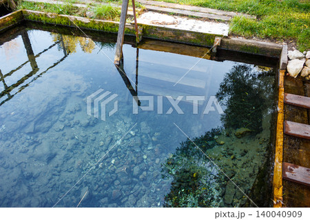 Clear, pure spring water in a rustic wooden plunge pool. Visible rocks below, reflecting green foliage and wooden structures. Serene spot for a refreshing dip or baptismal rite Clear, pure spring water in a rustic wooden plunge pool. Visible rocks below, reflecting green foliage and wooden structures. Serene spot for a refreshing dip or baptismal rite 140040909