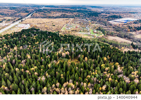 Sweeping aerial vista of a mixed autumn woodland, vibrant greens meeting golden hues, contrasting with a distant road and village dwellings 140040944