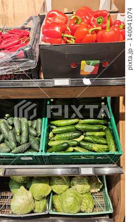 Fresh red bell peppers, cucumbers, and green cabbage displayed in crates on wooden shelves inside a grocery store produce section Fresh red bell peppers, cucumbers, and green cabbage displayed in crates on wooden shelves inside a grocery store produce section 140041074
