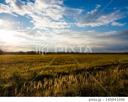 Vast golden field stretching under a dynamic sky with scattered clouds. Sunlight bathes the autumn grasses, creating a serene, expansive rural scene at dusk Vast golden field stretching under a dynamic sky with scattered clouds. Sunlight bathes the autumn grasses, creating a serene, expansive rural scene at dusk 140041096