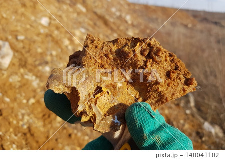 A gloved hand cradles a rough, earthy piece of opaque chalcedony on flint, showcasing its rugged texture against a soft-focus barren landscape under bright sunlight A gloved hand cradles a rough, earthy piece of opaque chalcedony on flint, showcasing its rugged texture against a soft-focus barren landscape under bright sunlight 140041102