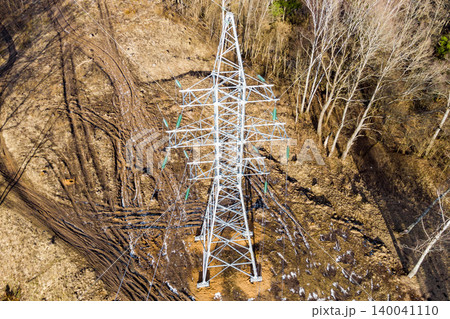 Towering electricity pylon stands on rugged ground with tire tracks. Bare trees flank the scene, hinting at a raw, untamed landscape. Utility and nature intertwine 140041110