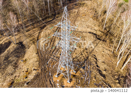 Aerial view of a massive power pylon amidst a rugged, early spring landscape with sparse trees and dry ground, symbolizing energy transmission 140041112